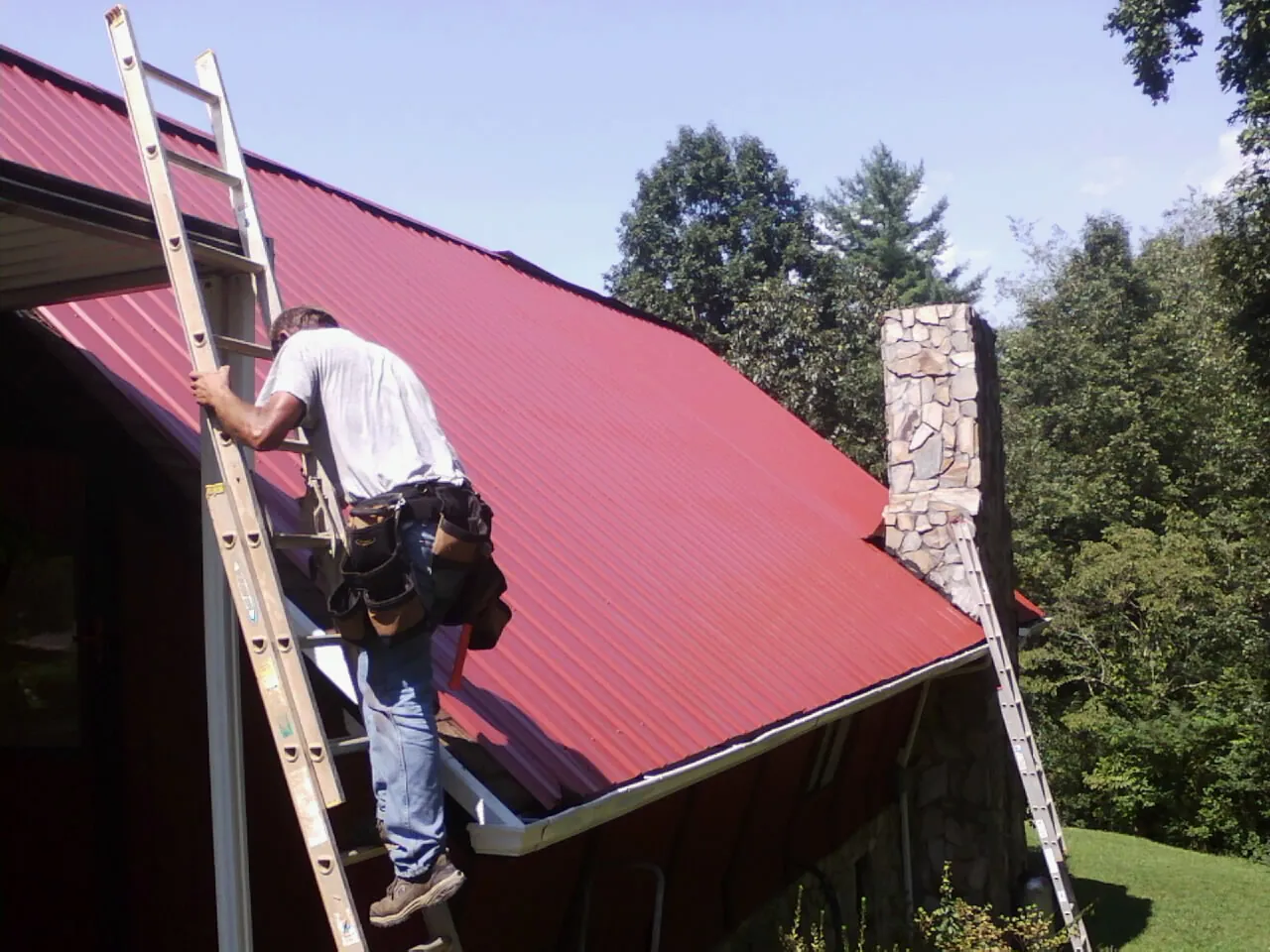 Roofer on a ladder beside a red metal roof and stone chimney — Soffit & Fascia Repair by Roofing Brothers in Lapaz